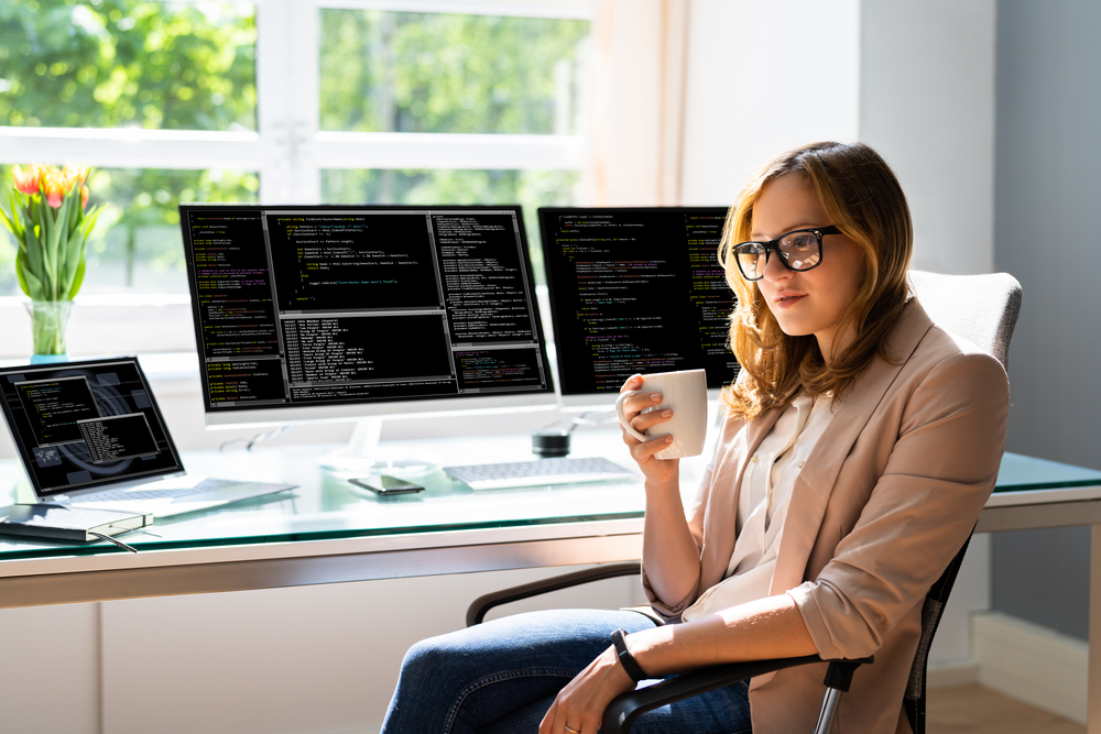 A blonde woman with glasses, wearing jeans and a tan blazer, sitting at a desk with two monitors and a laptop in a well lit room with gerber daisies in the window. She is drinking a cup of coffee.